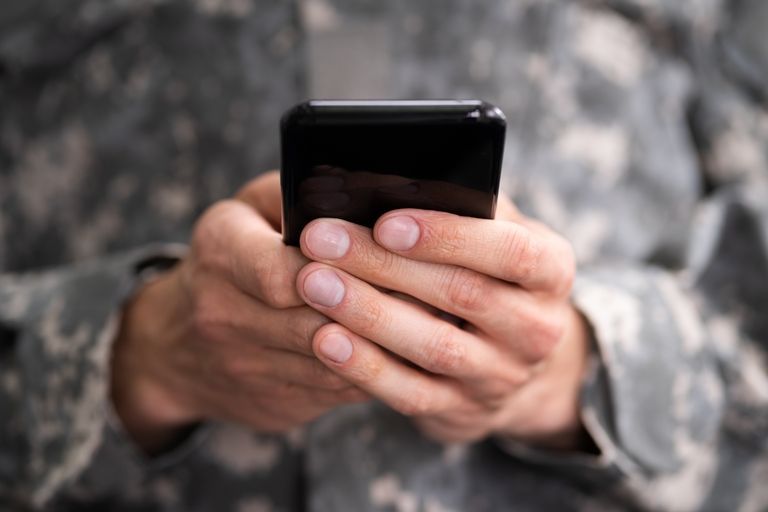 Military personnel is holding a cell phone in his hands.