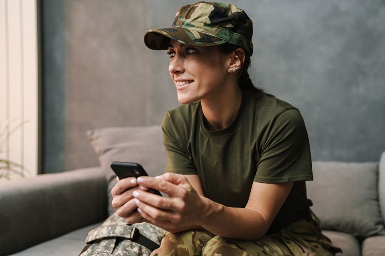 A woman in a military uniform is sitting on a couch using a cell phone.
