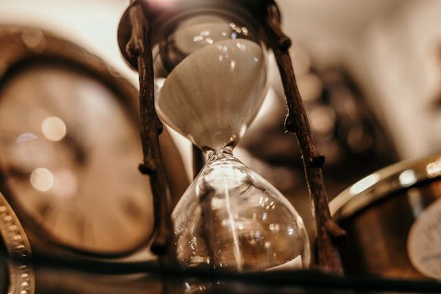 Hourglass with sand flowing, set amongst other antique clocks. Warm lighting.