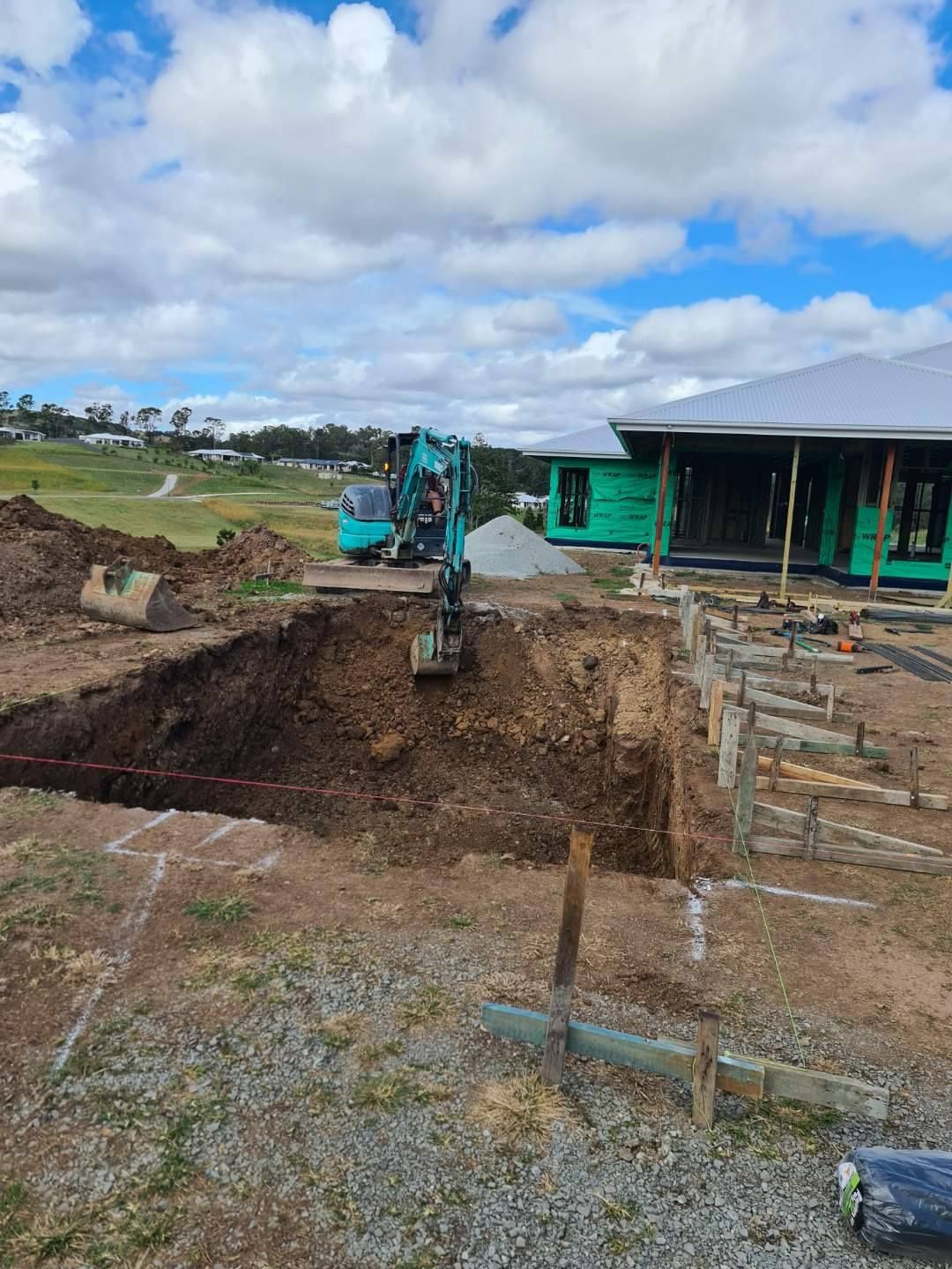 An excavator is digging a hole in the ground in front of a house under construction — CJB Pools in Gympie, QLD