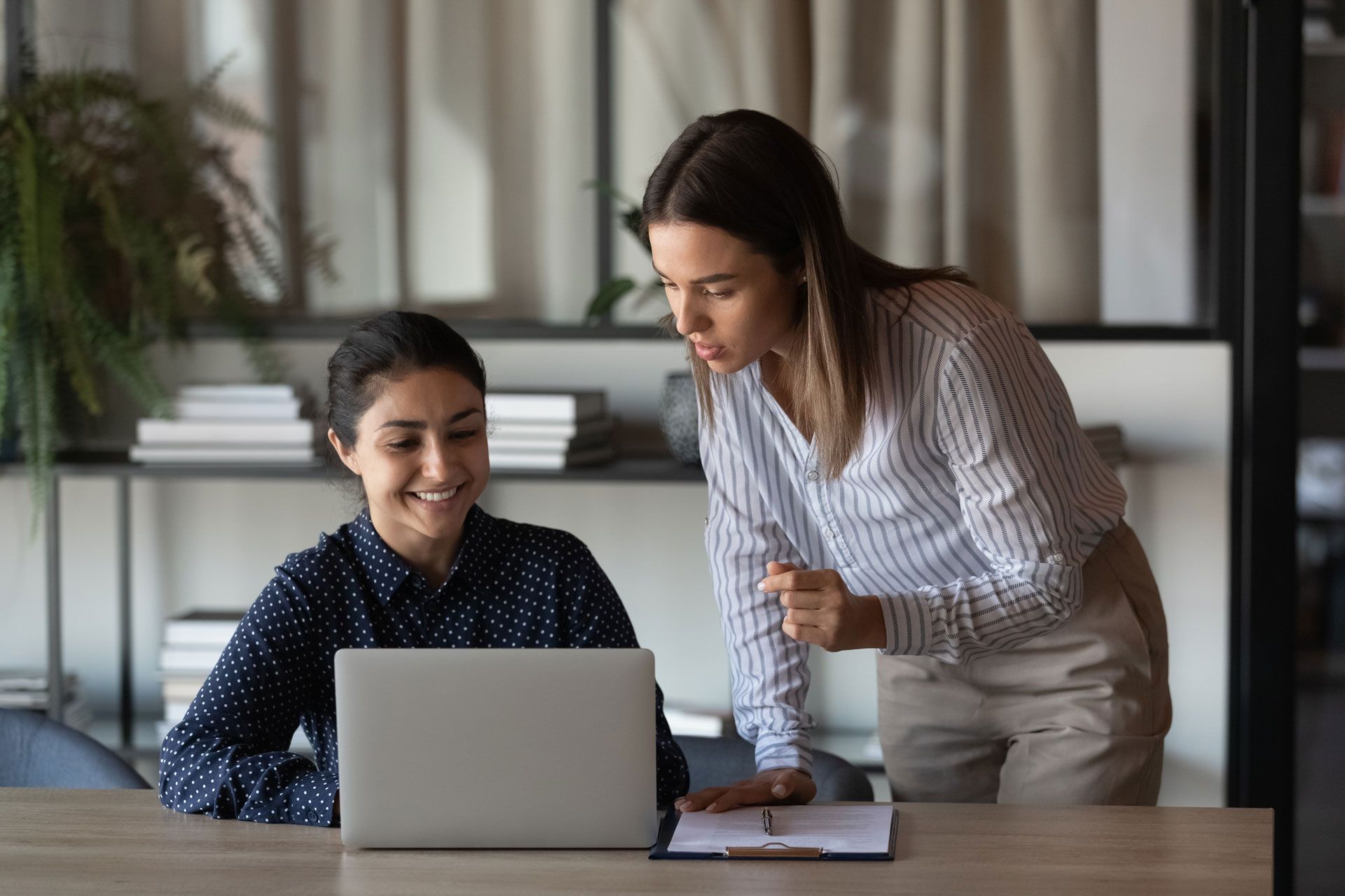 Two women looking at a laptop in an office. One is seated, smiling; the other is standing, pointing.