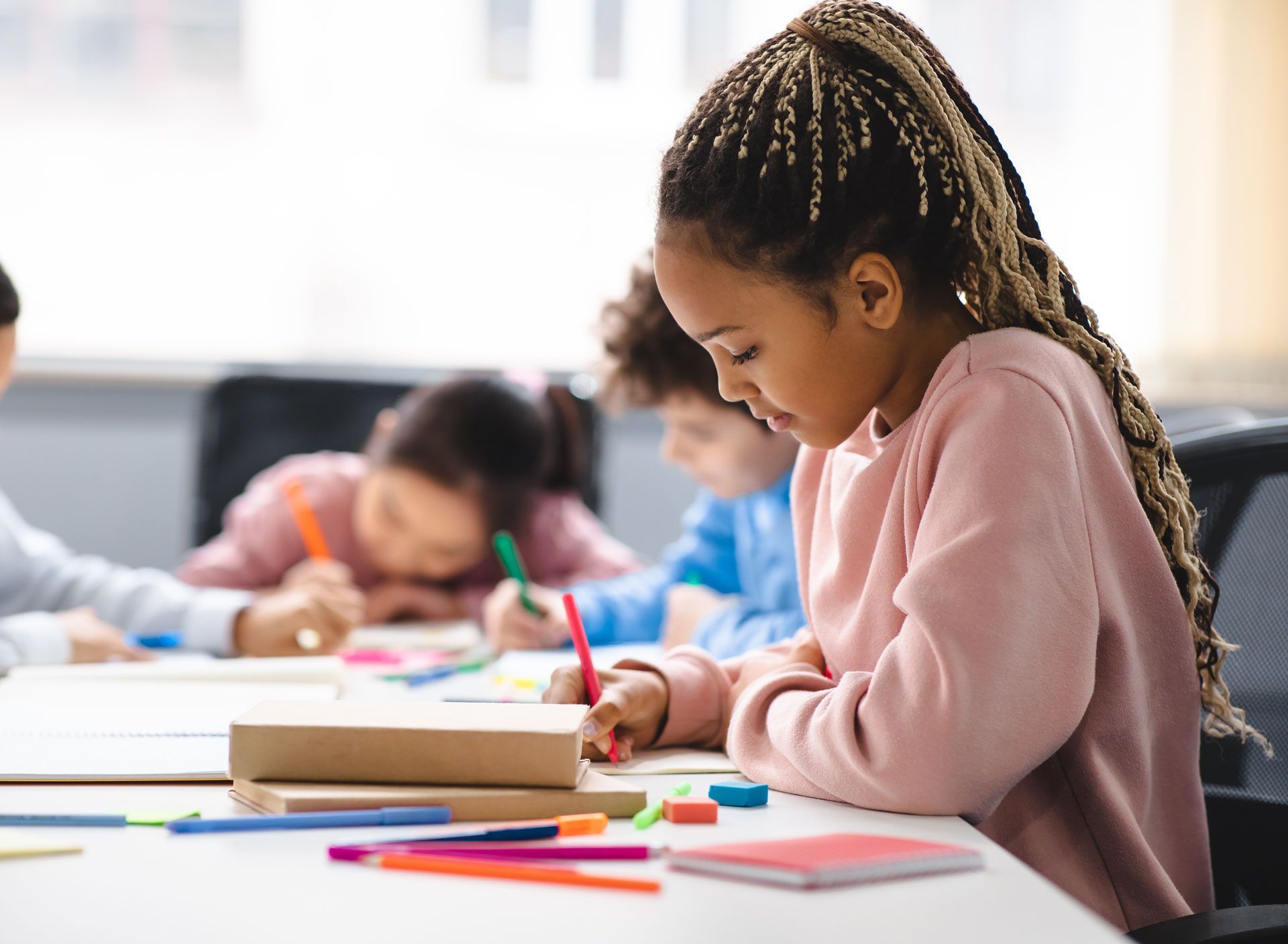 A young Black girl with braided hair, focused on drawing at a table with other children in a classroom.