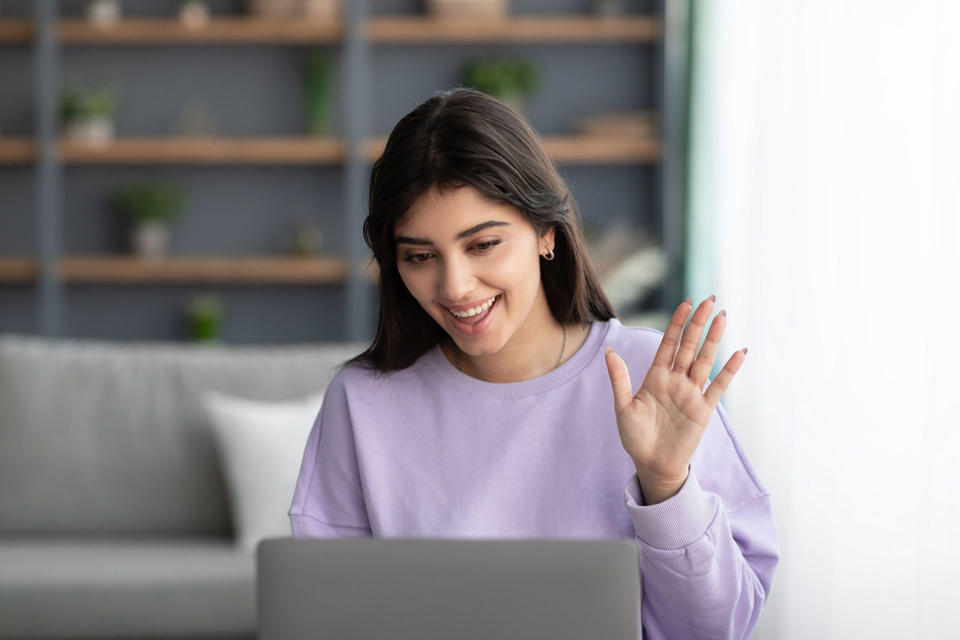 Woman in a purple sweater waves at a laptop, smiling. She's indoors near a sofa and shelves.