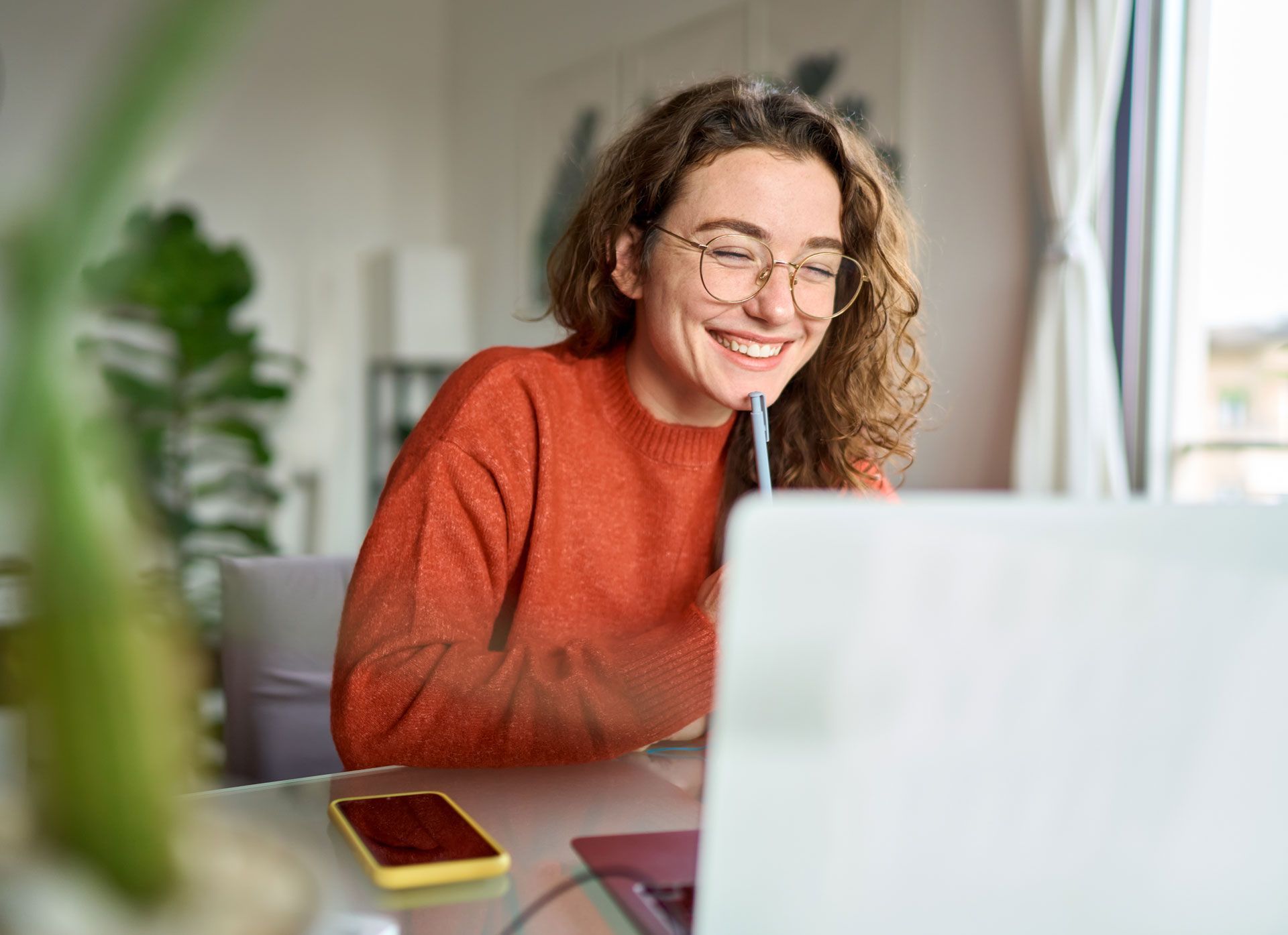 Woman with curly hair, glasses, and orange sweater smiles while looking at a laptop in a bright room. She is holding a pen.