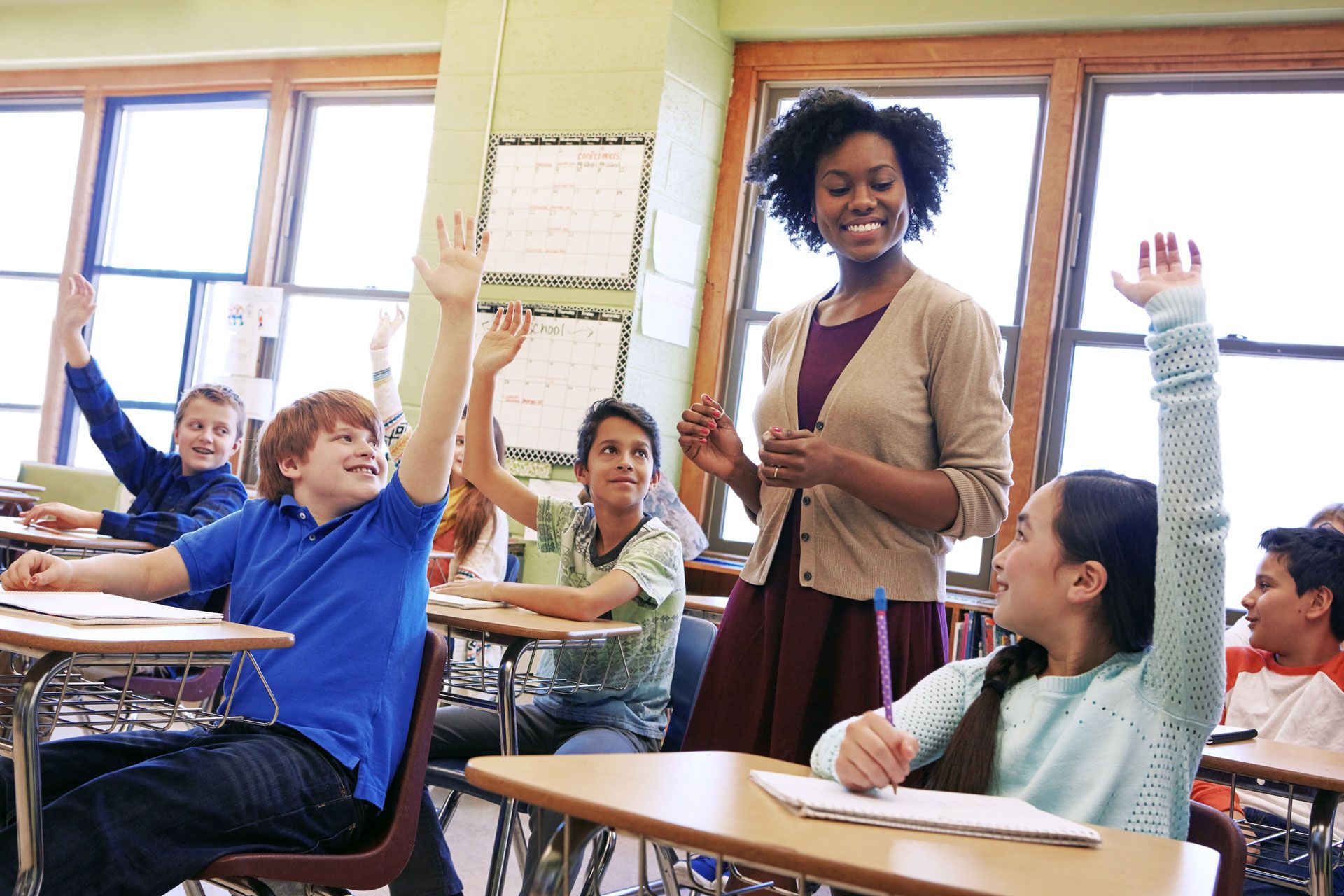 Teacher smiles at students with raised hands in a brightly lit classroom.