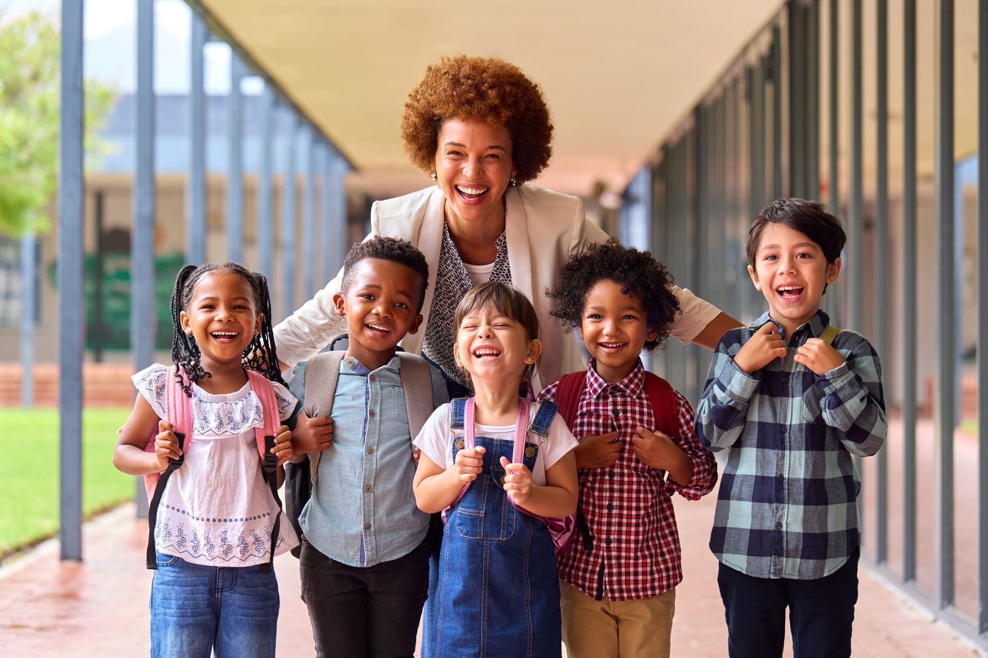 Teacher smiling with group of children wearing backpacks, standing outside a school.