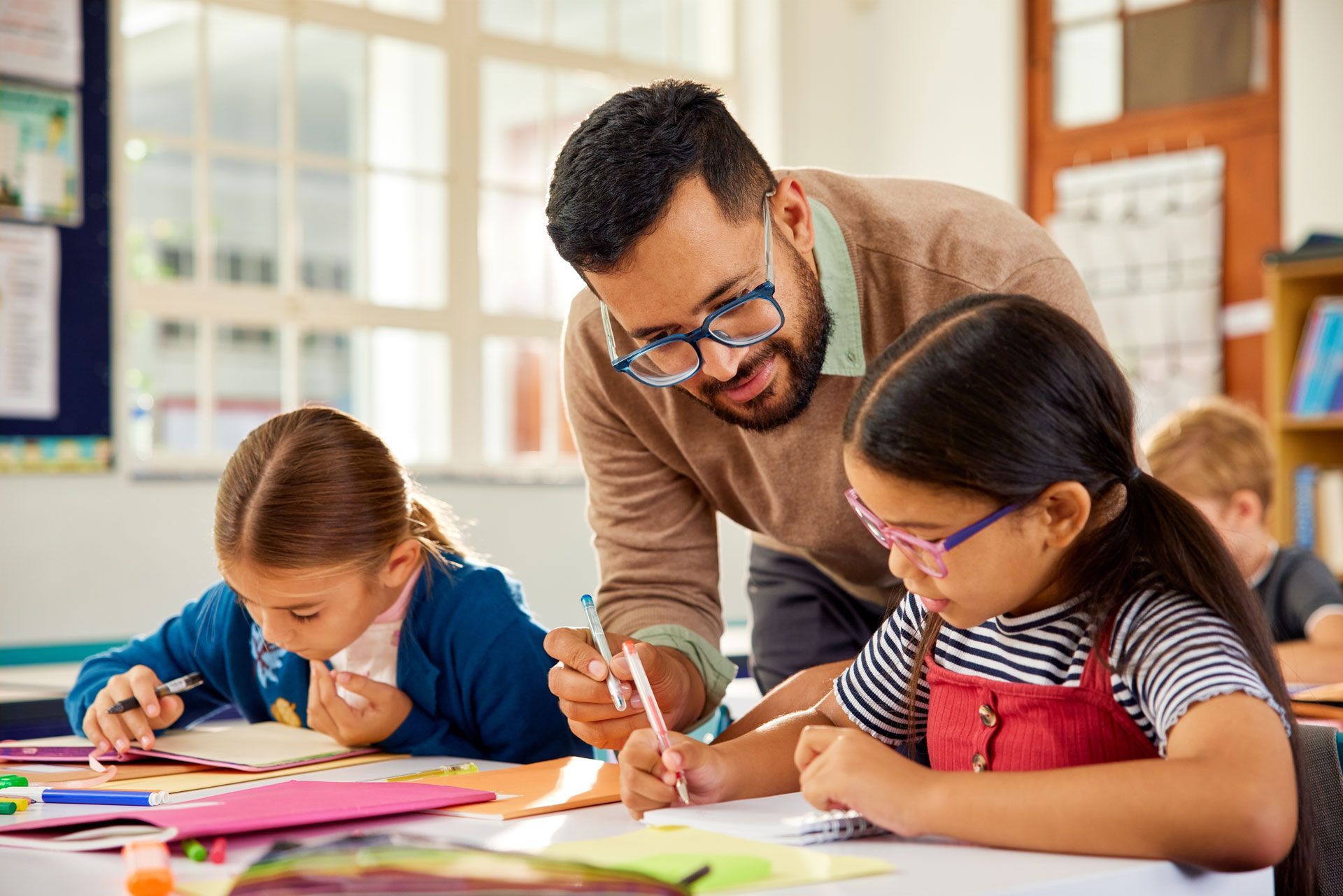 Teacher assisting a student with writing at a desk, another student nearby. Classroom setting.