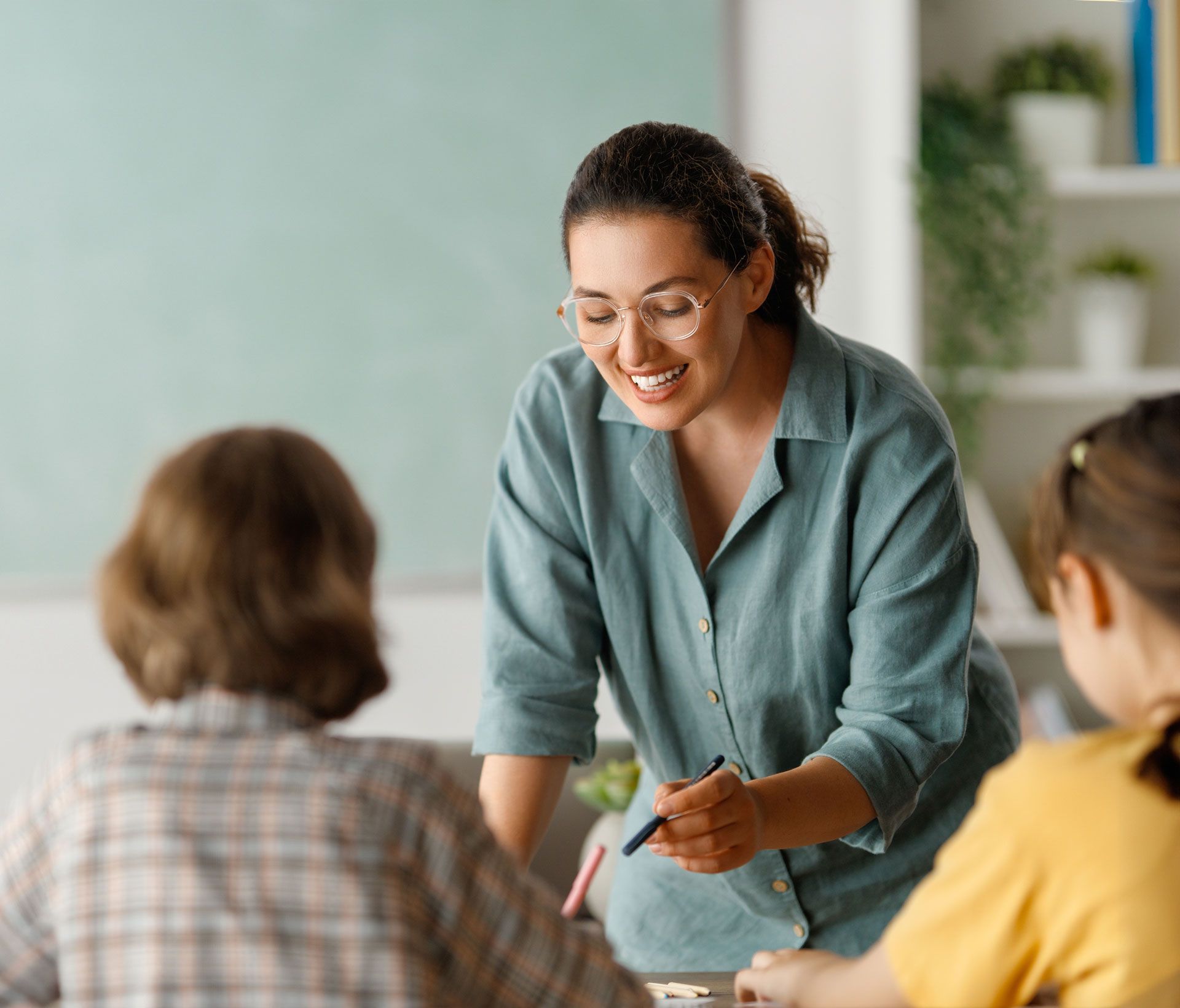 Teacher smiling, leaning over students at a table in a classroom, pointing a pen.