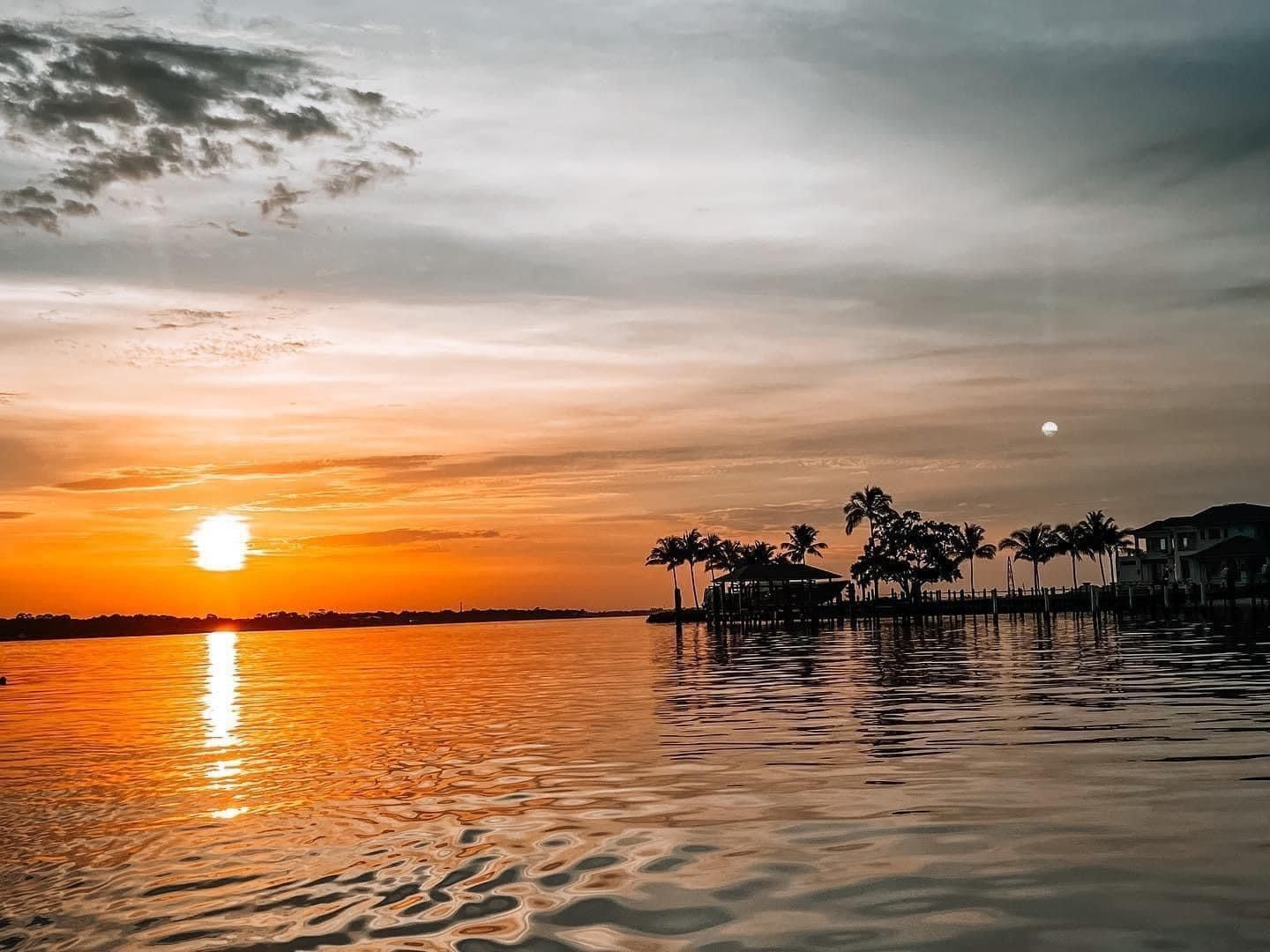 The sun is setting over a body of water with palm trees in the foreground.