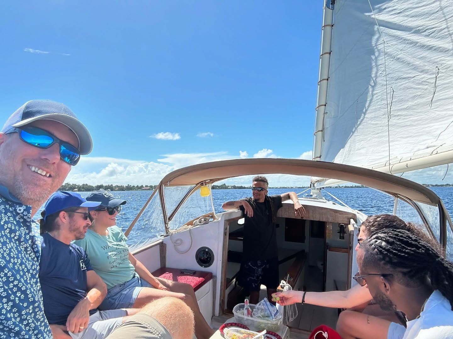 People sailing on a boat on a sunny day; one man takes a selfie, others relax, blue water and sky.