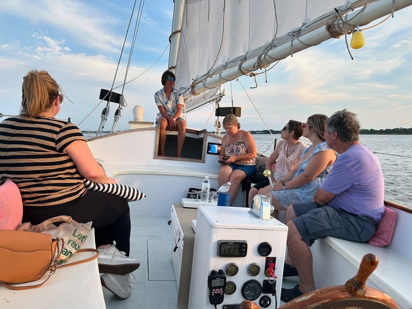 People on sailboat; woman sitting, others seated on bench, water and sky visible.