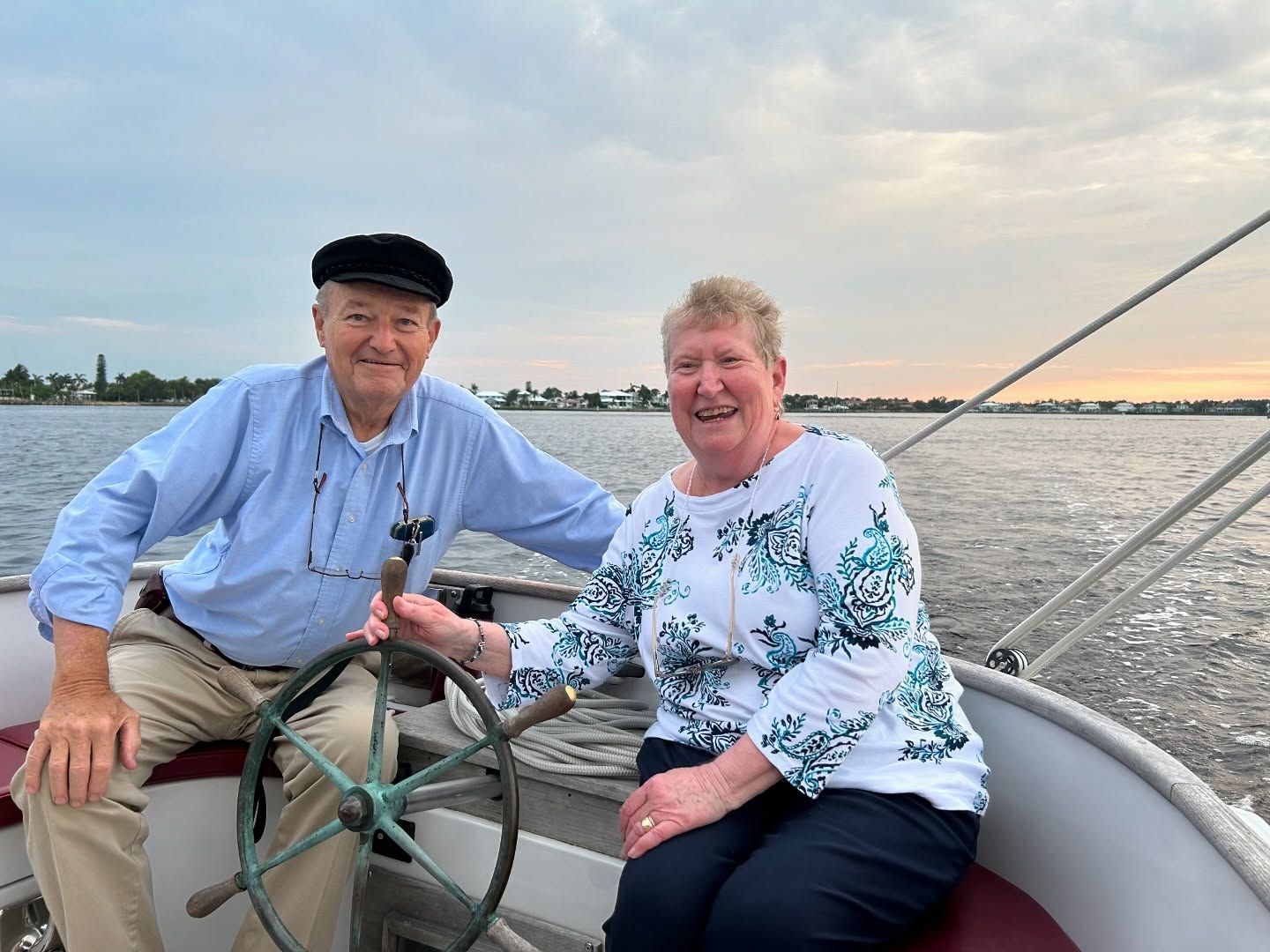 Older couple on a boat, man at the wheel, woman next to him, smiling, cloudy sky over water.