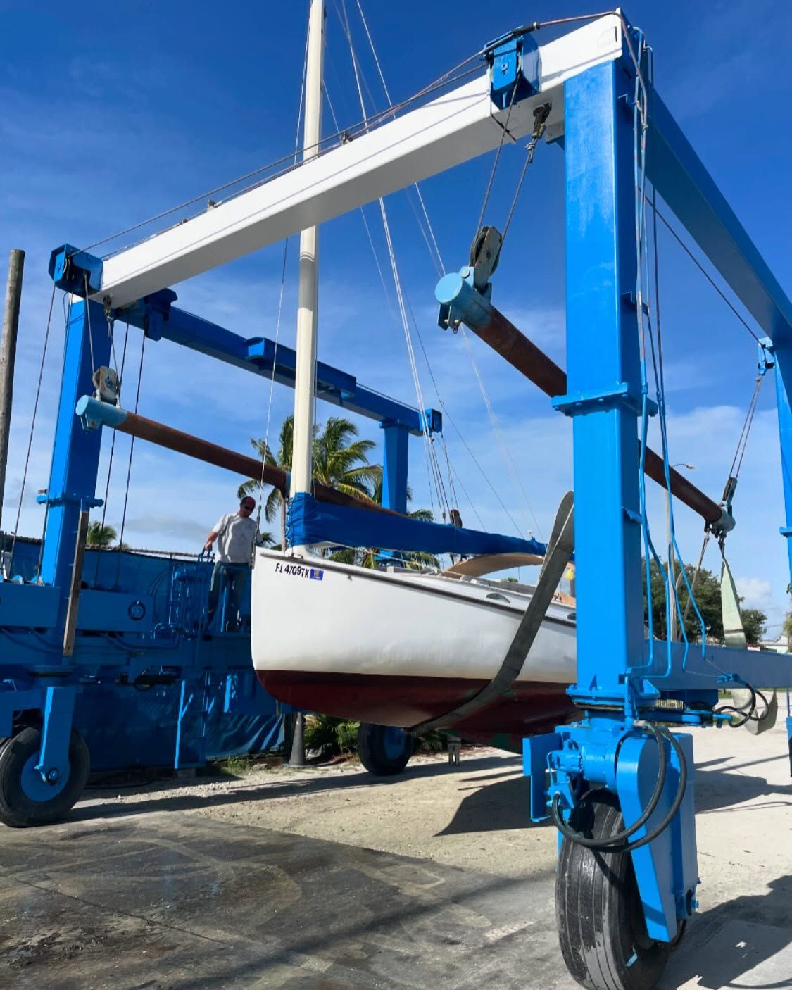 Sailboat being lifted by a blue and white boat lift, with a man standing nearby under a blue sky.