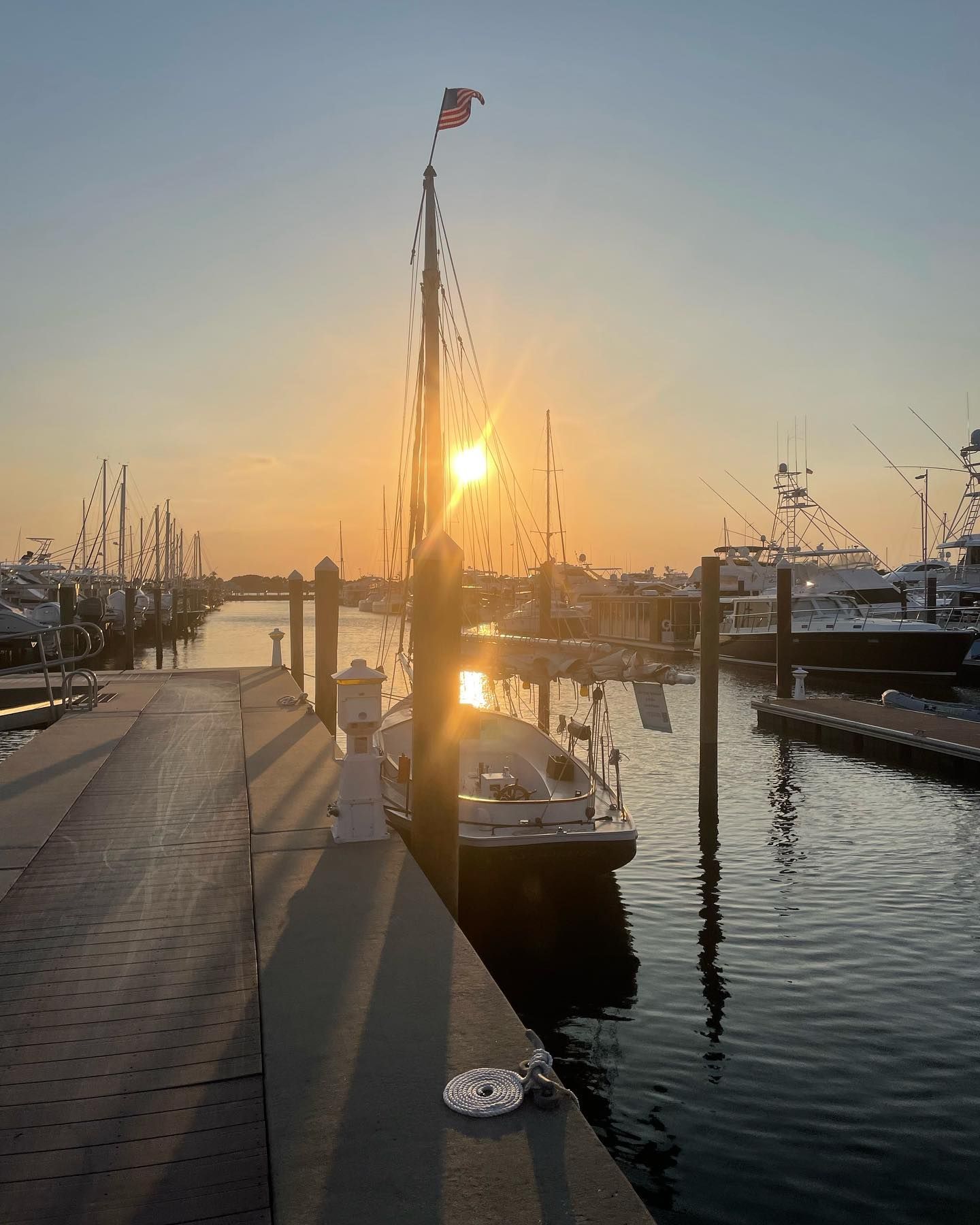 Sunset at a marina, a sailboat is docked. Sun shines behind the mast. Water reflects the warm colors.