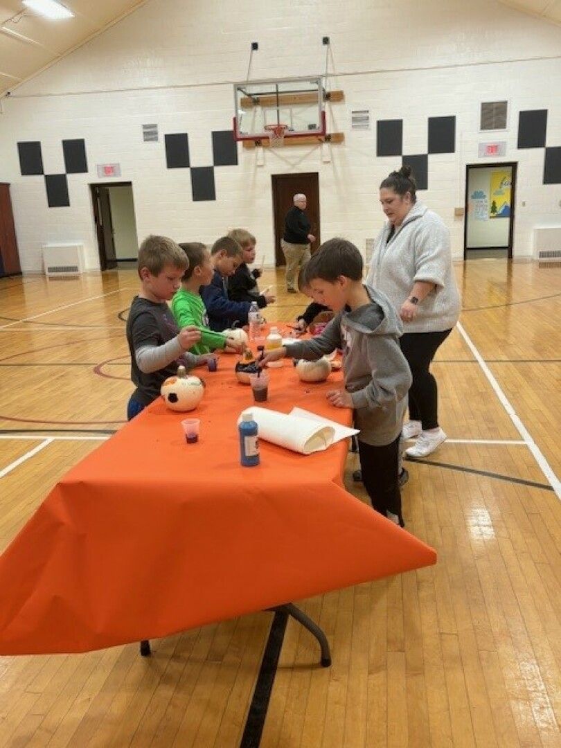 Children enthusiastically paint small pumpkins at an orange-covered table in a gym. 