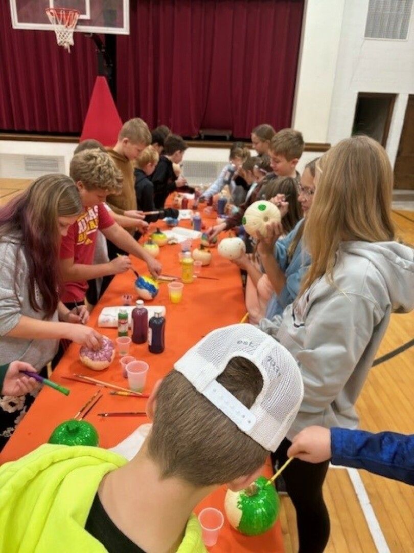 A group of children in a gymnasium engage in painting small pumpkins at a table covered with an orange cloth.