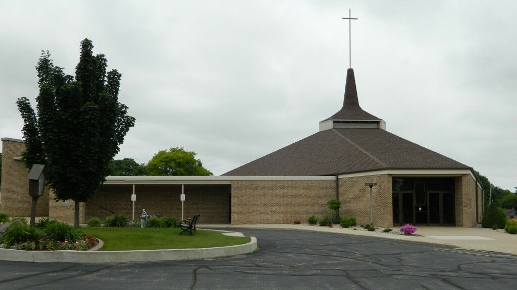 A modern brick church with a tall steeple topped by a cross stands against a cloudy sky. 