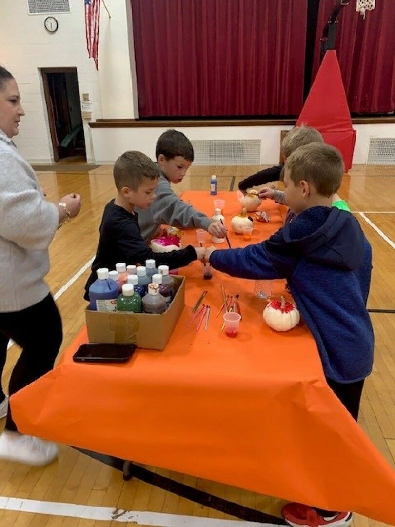Children are painting small pumpkins at a table covered with an orange cloth in a gym.