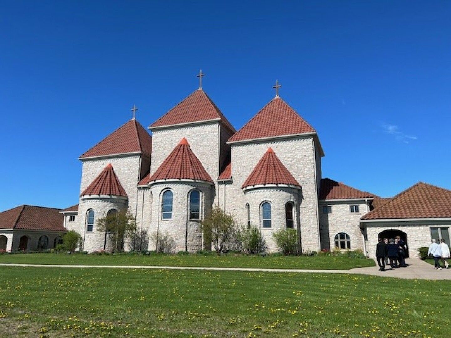 A large church with red-tiled, conical roofs and stone walls stands against a clear blue sky. 