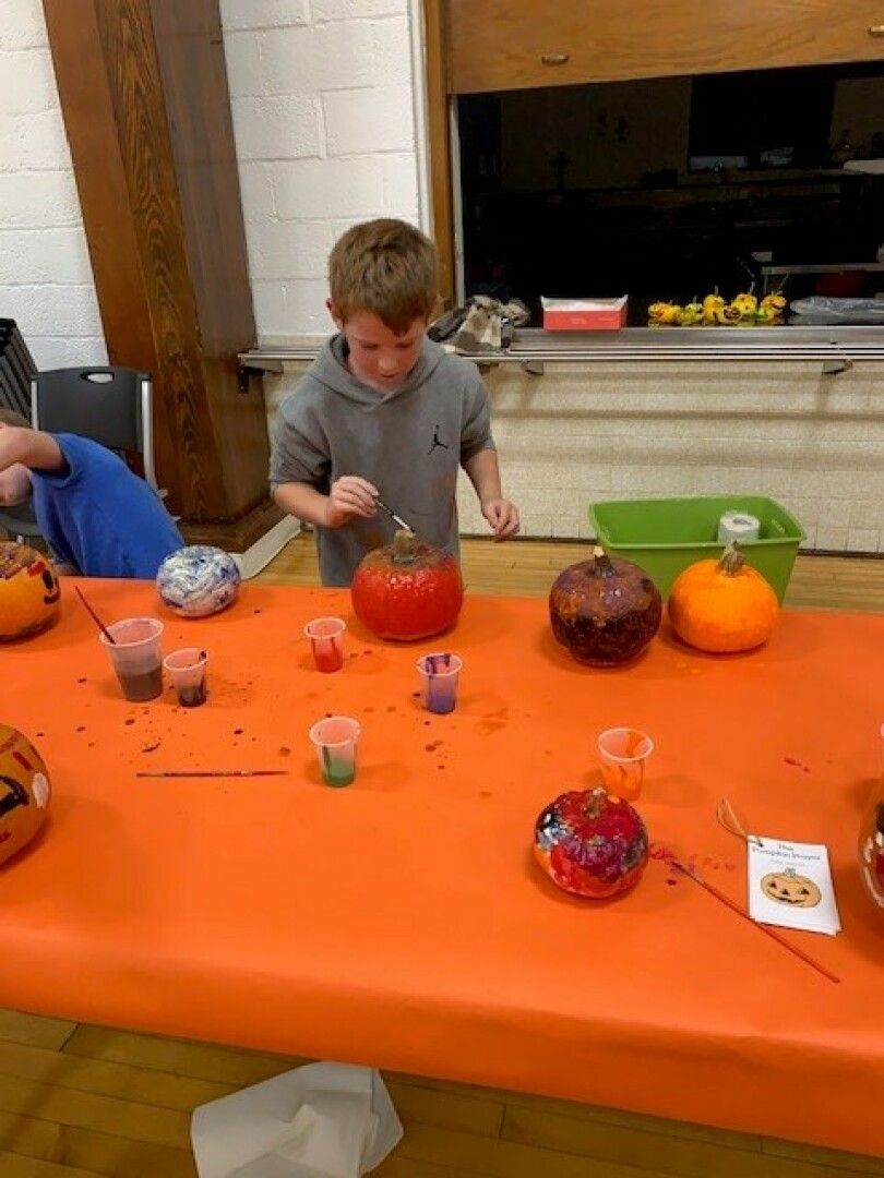 A young boy paints a pumpkin at a craft table covered with an orange cloth.
