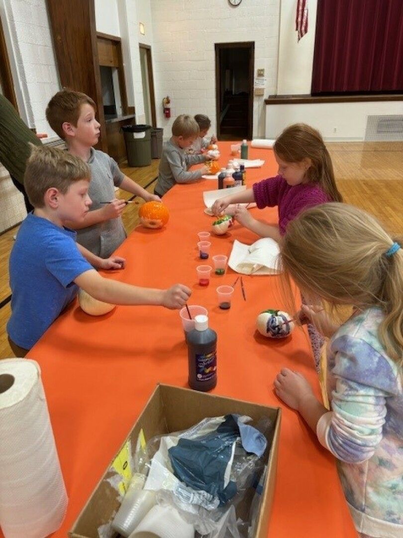 Children are gathered around an orange table, painting small pumpkins. 