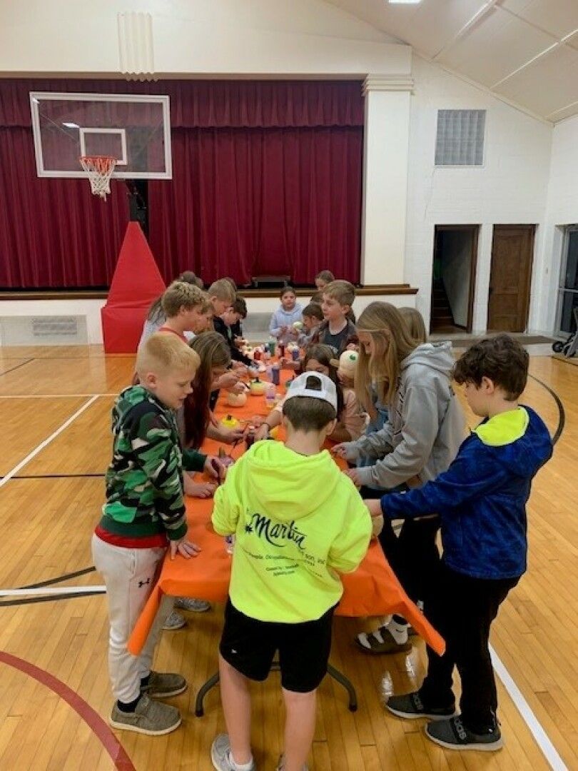 A group of children and teens are gathered around a craft table in a gymnasium.