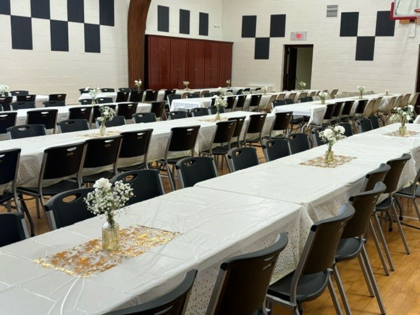 A banquet hall with long tables covered in white cloths, adorned with small vases of white flowers and black chairs.