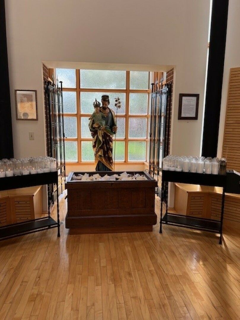 A wooden statue of a saint stands by a sunlit window, flanked by lit white candles on black metal stands.