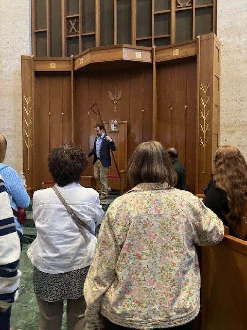 A man in a blazer speaks to a group in a wood-paneled room with tall geometric designs. 
