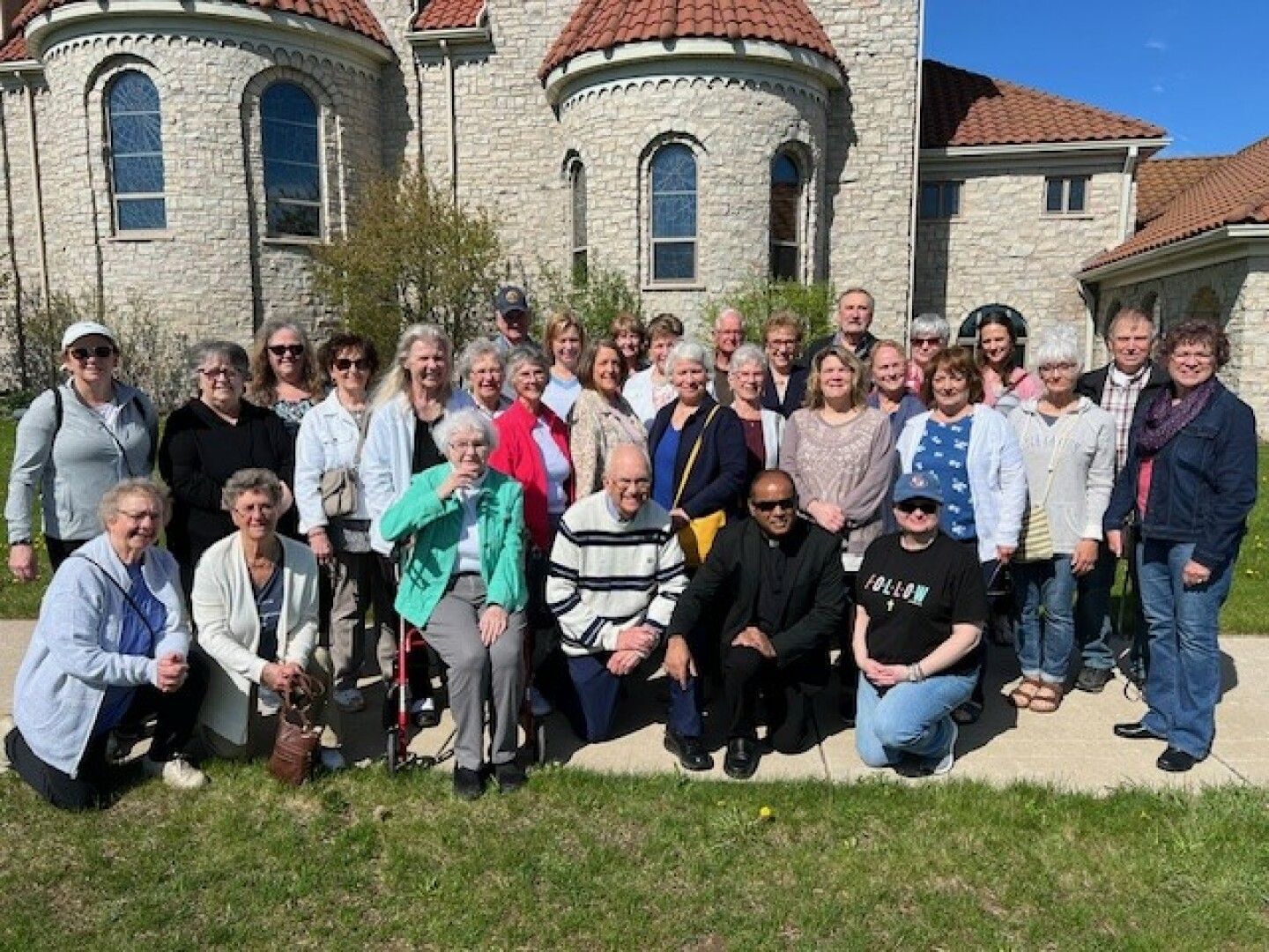 A large group of smiling people poses outside a stone building with arched windows and red-tiled roofs. 