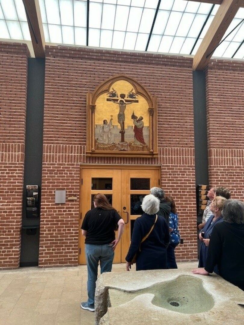 A group of people admire a religious artwork depicting the crucifixion, mounted on a brick wall under a glass ceiling.