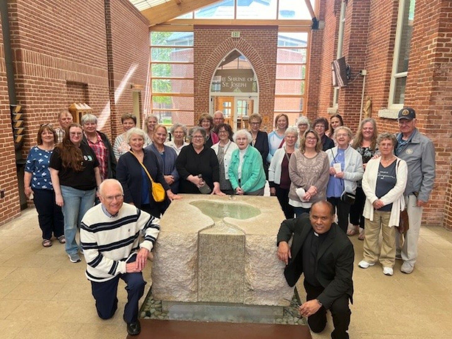 A group of around 25 smiling people, mostly older adults, gathered inside a brick building with a large stone baptismal font. 
