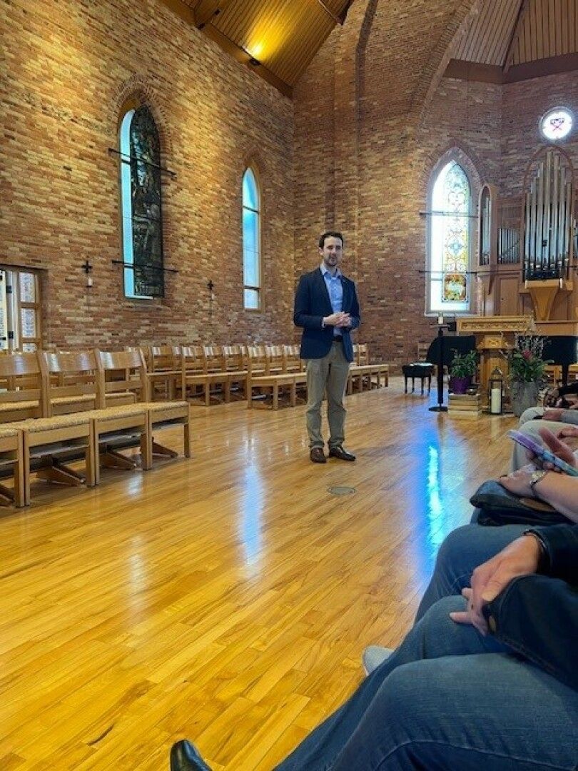Man in formal attire stands in a serene chapel setting with brick walls and wooden floor, speaking to seated audience. 
