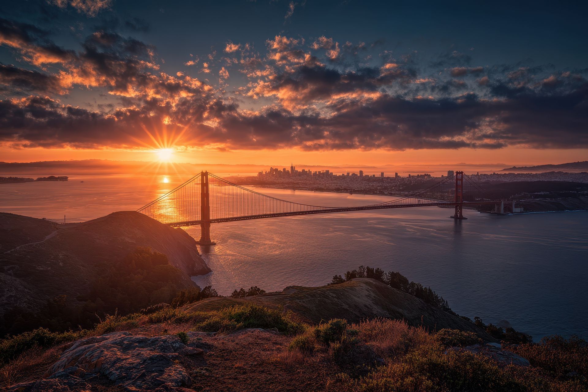 Golden Gate Bridge at sunset, sun reflecting on water, San Francisco skyline in background, dramatic orange sky.