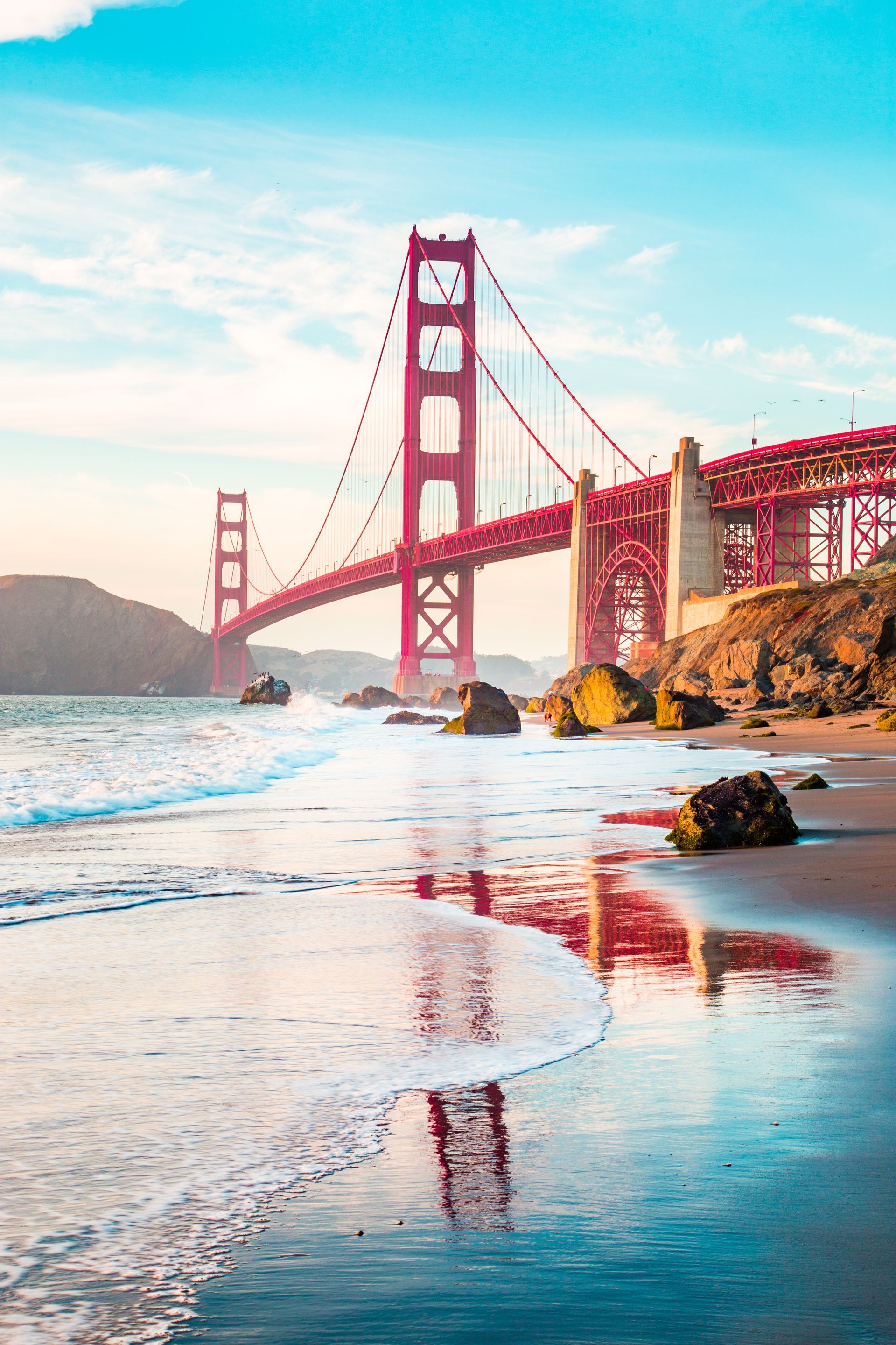 Golden Gate Bridge, red, over water. Blue sky, beach with rocks.