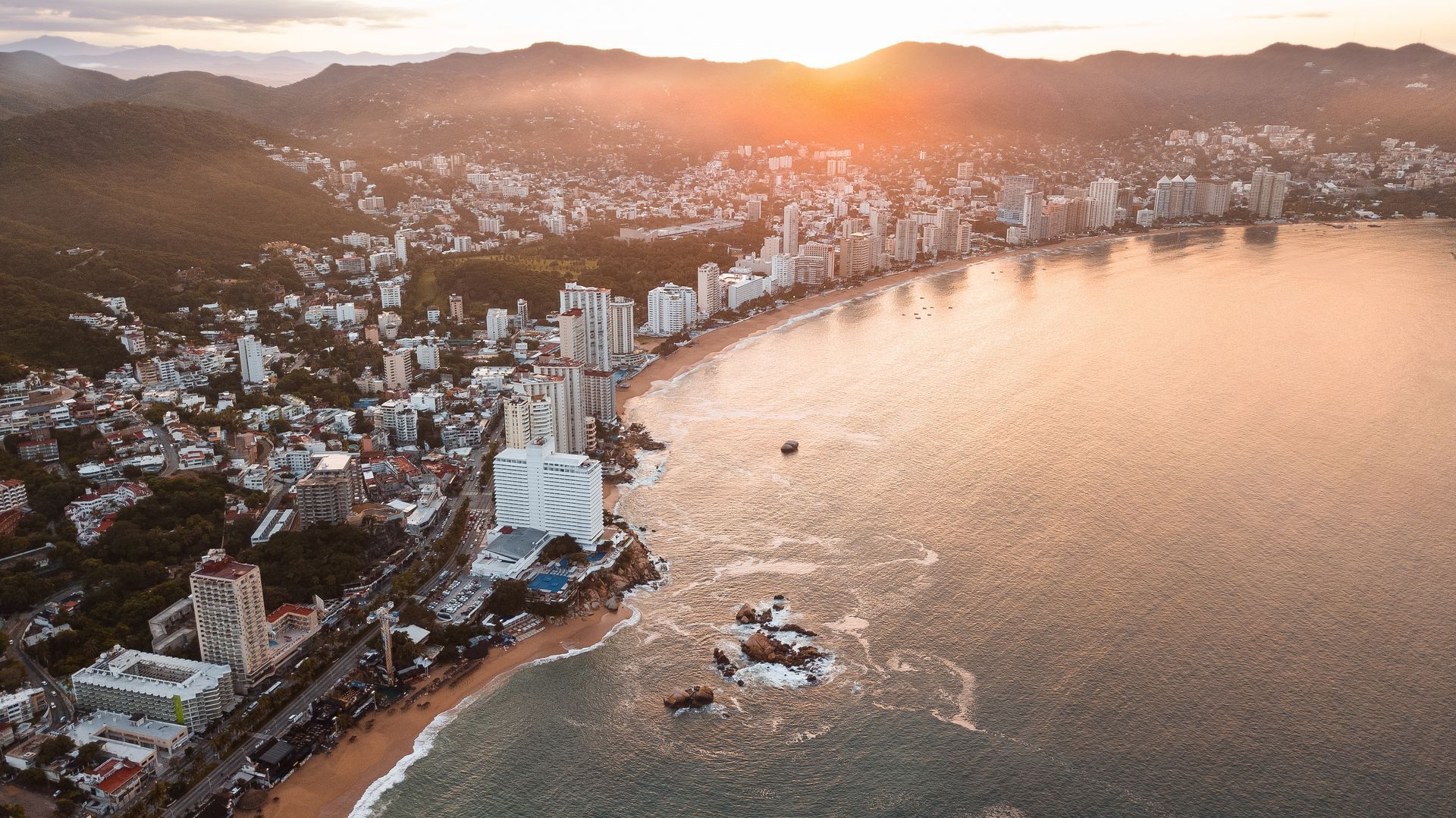 Coastal city at sunset, buildings line a bay. Warm sunlight reflects on the water.