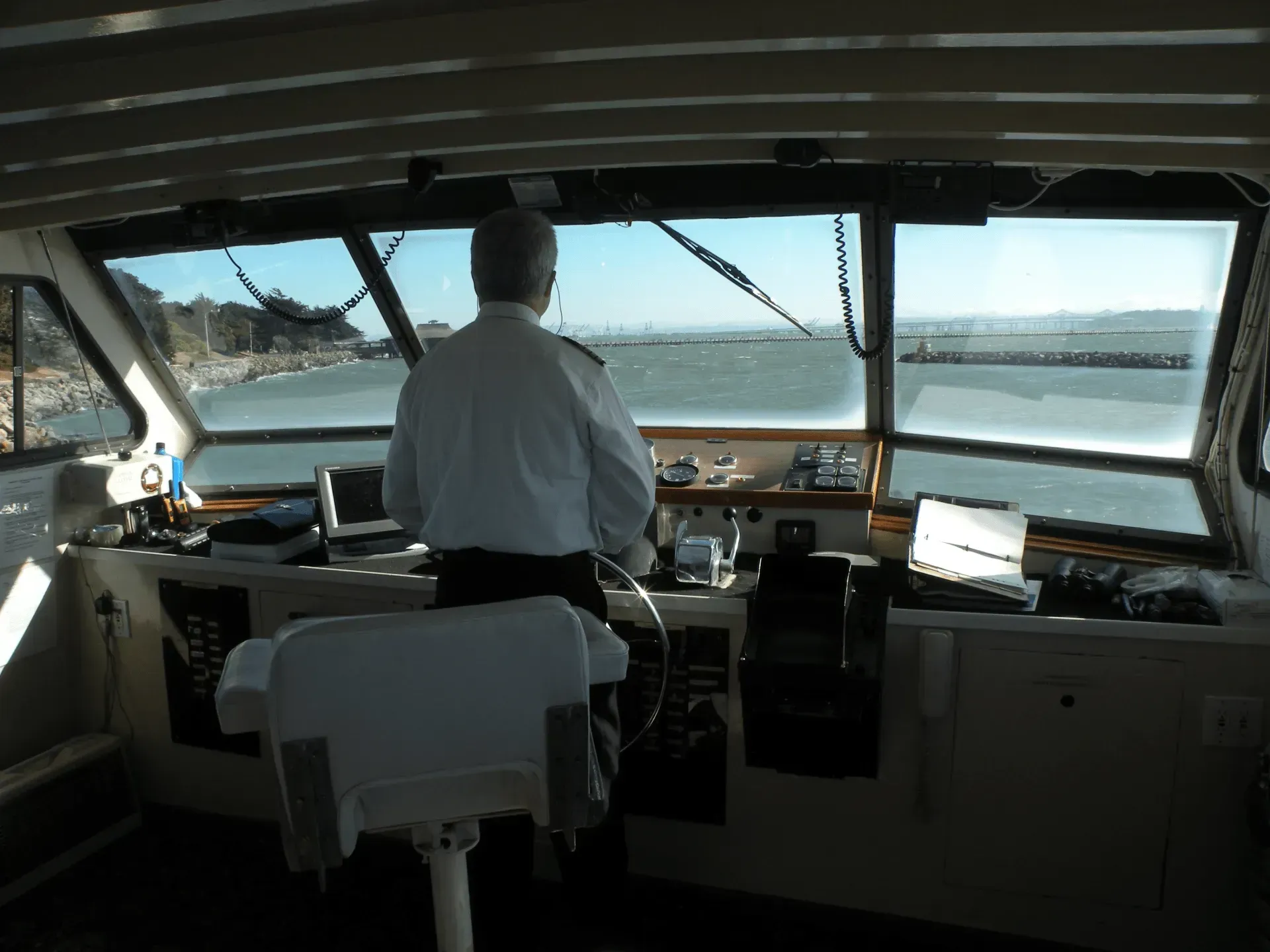 Person at the helm of a boat, looking out at the water. Inside the boat's bridge, bright daylight.