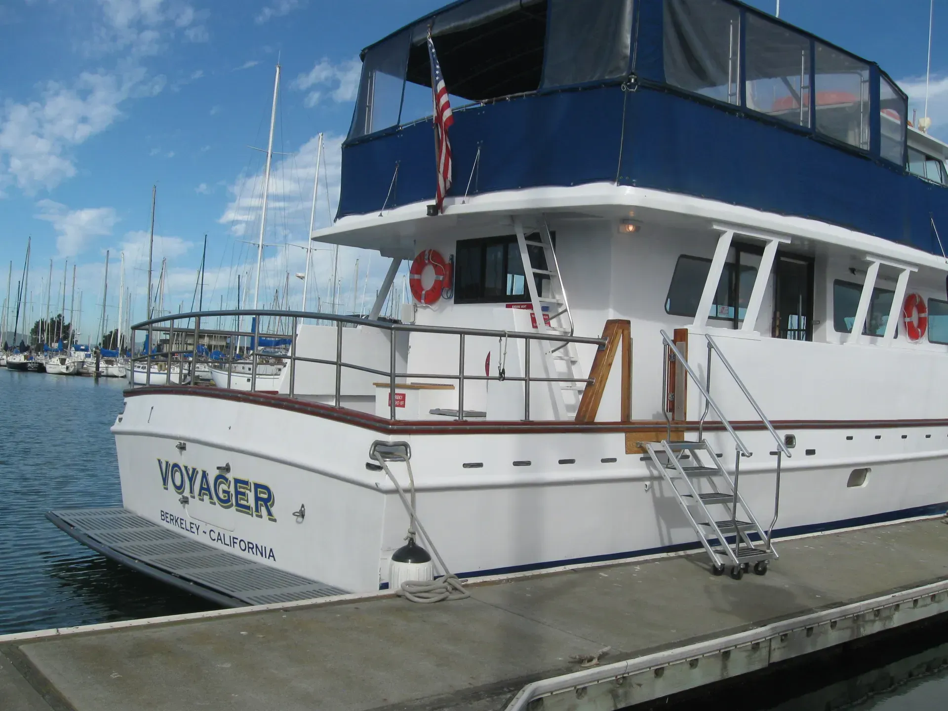 White boat Voyager at a marina, with boarding stairs and blue awning, an American flag flying.