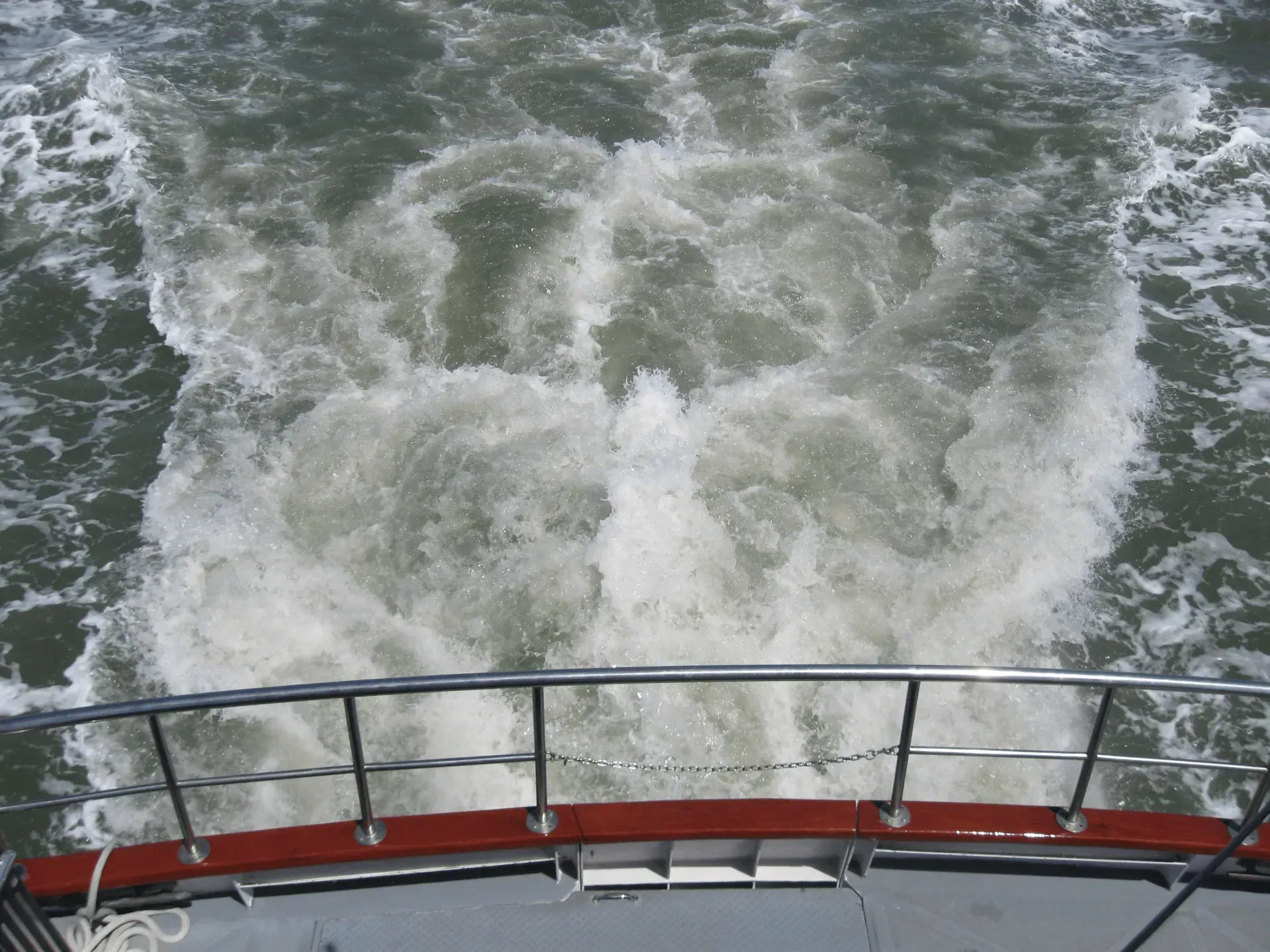 Boat's stern with white foamy wake on dark water. Stainless steel railing and boat deck in view.