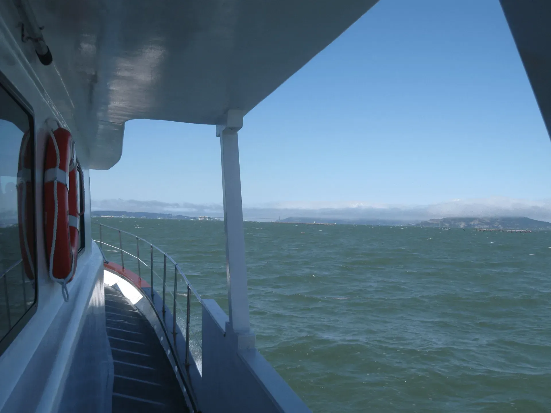 View from a boat deck, blue water, sky, life preserver on side of the vessel, distant shoreline.