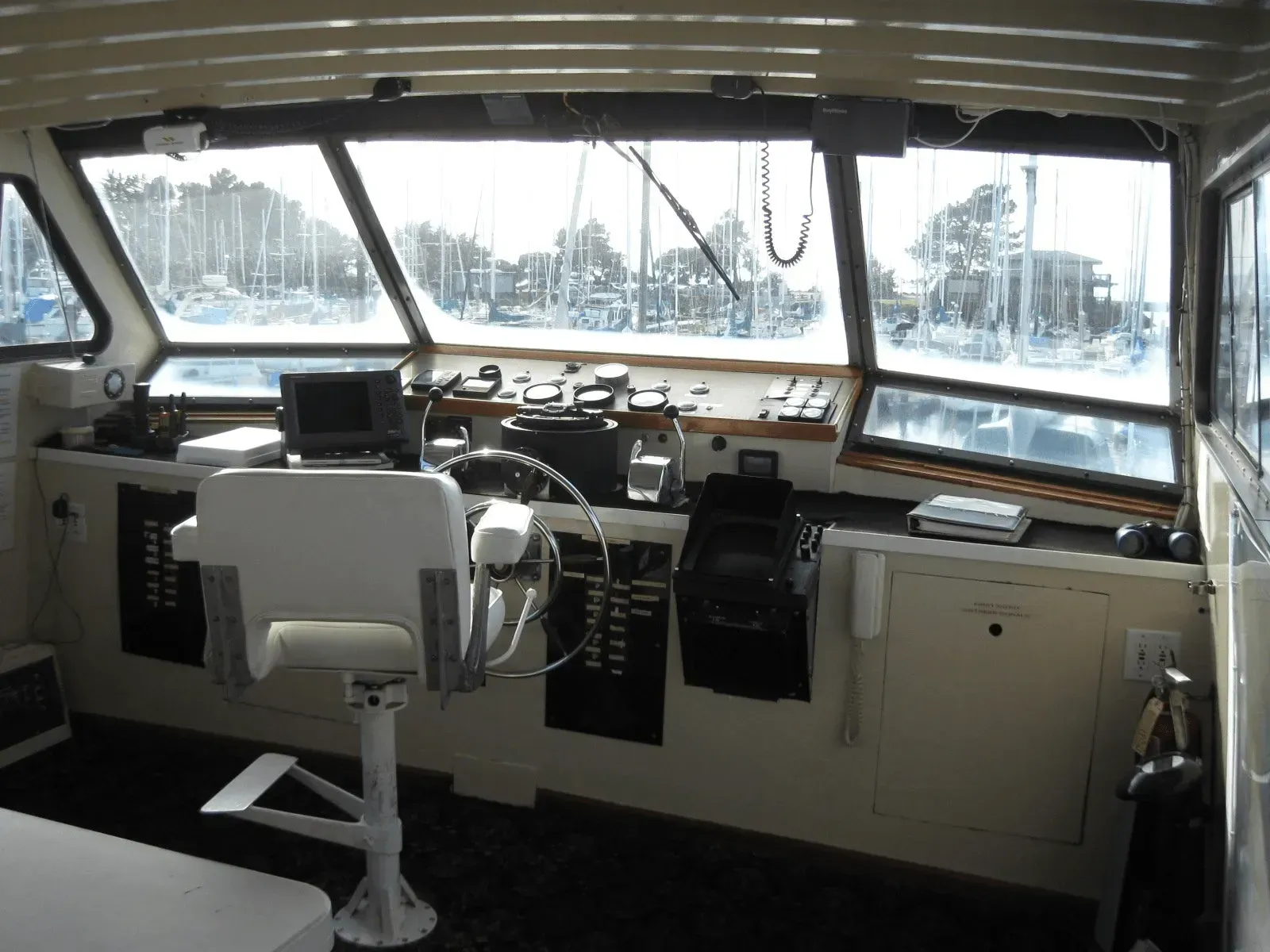 Interior view of a boat's navigation area with steering wheel, gauges, and large windows overlooking a marina.
