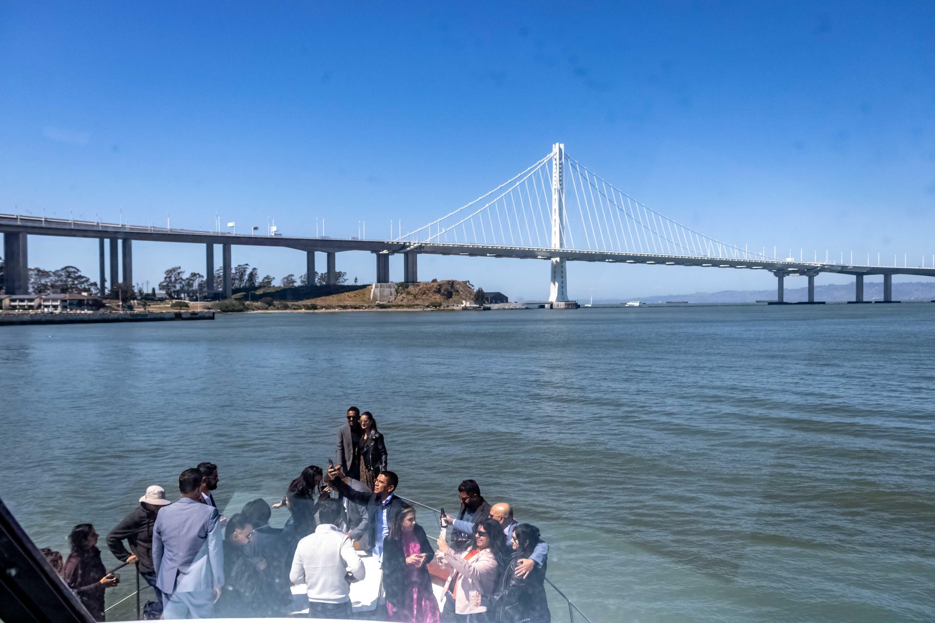 Friends on a boat at sunset, holding drinks, smiling. String lights and a bridge in the background.