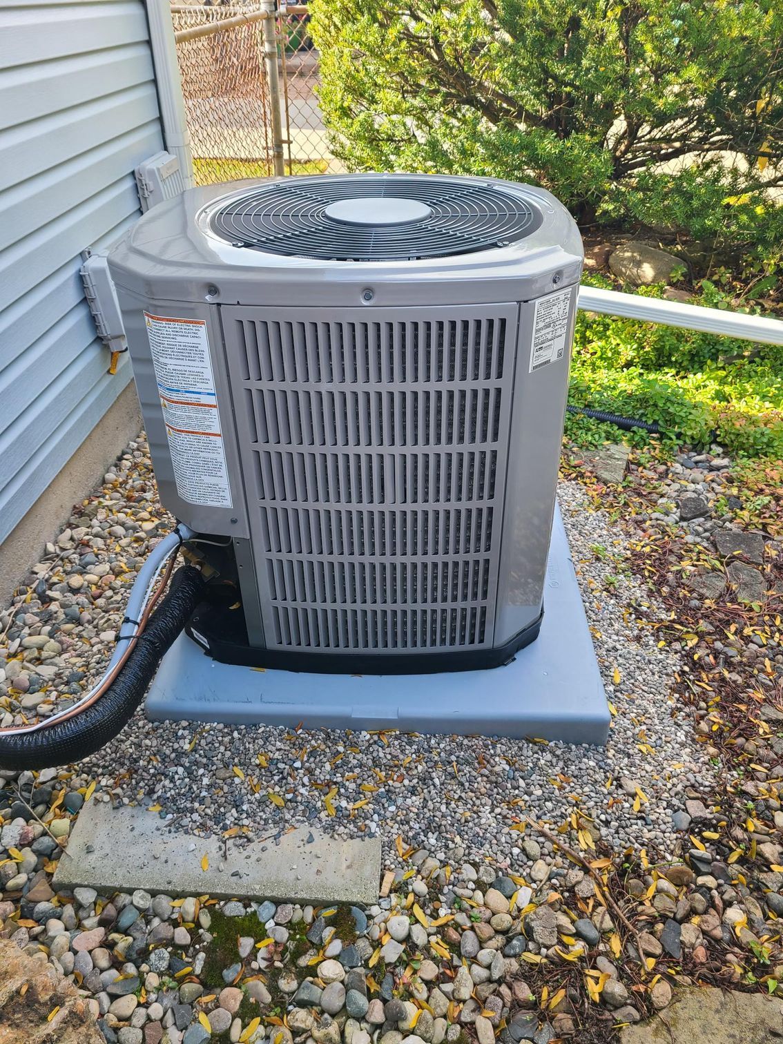 A gray air conditioner is sitting on top of a gravel area next to a house.