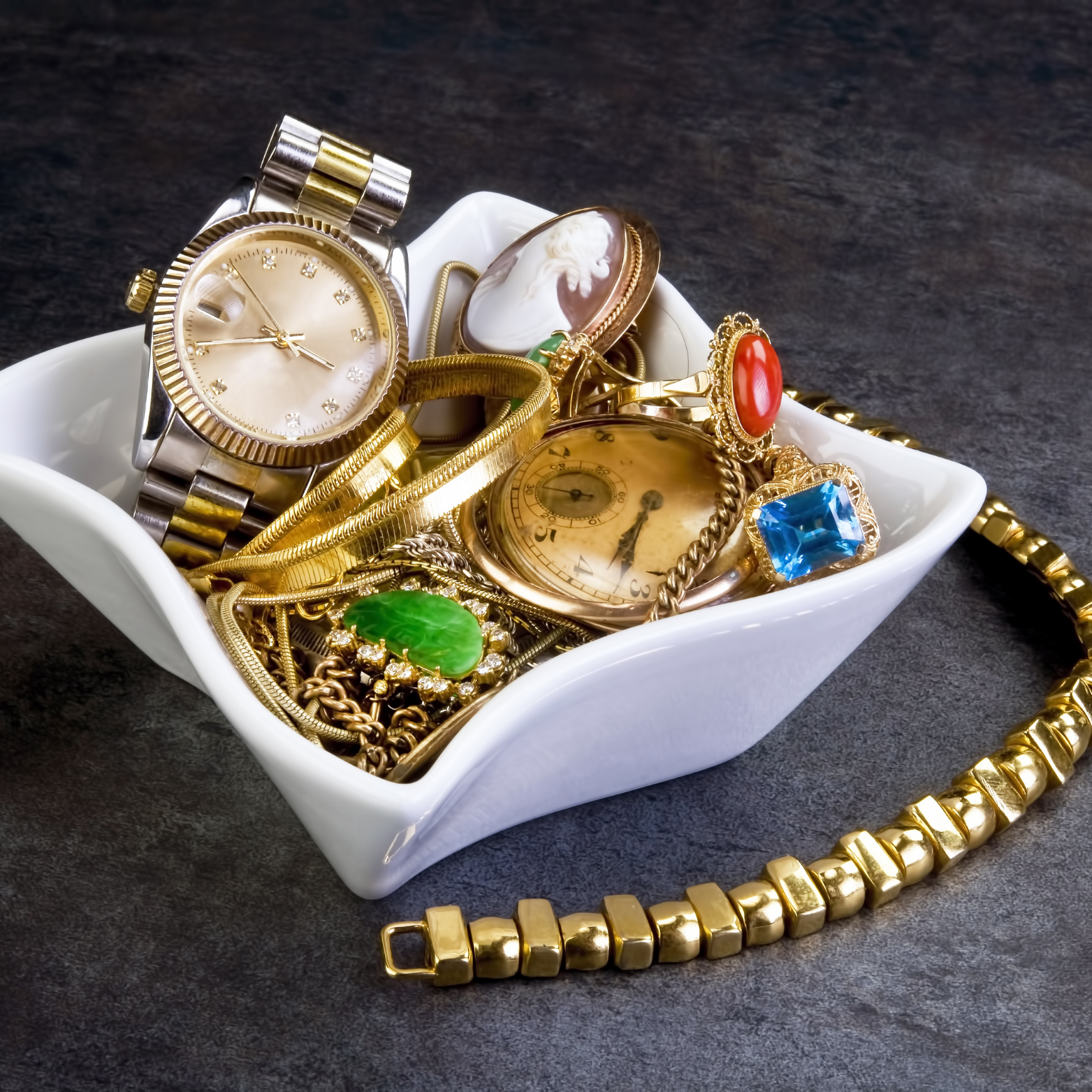 A white bowl filled with gold jewelry including a watch and a bracelet