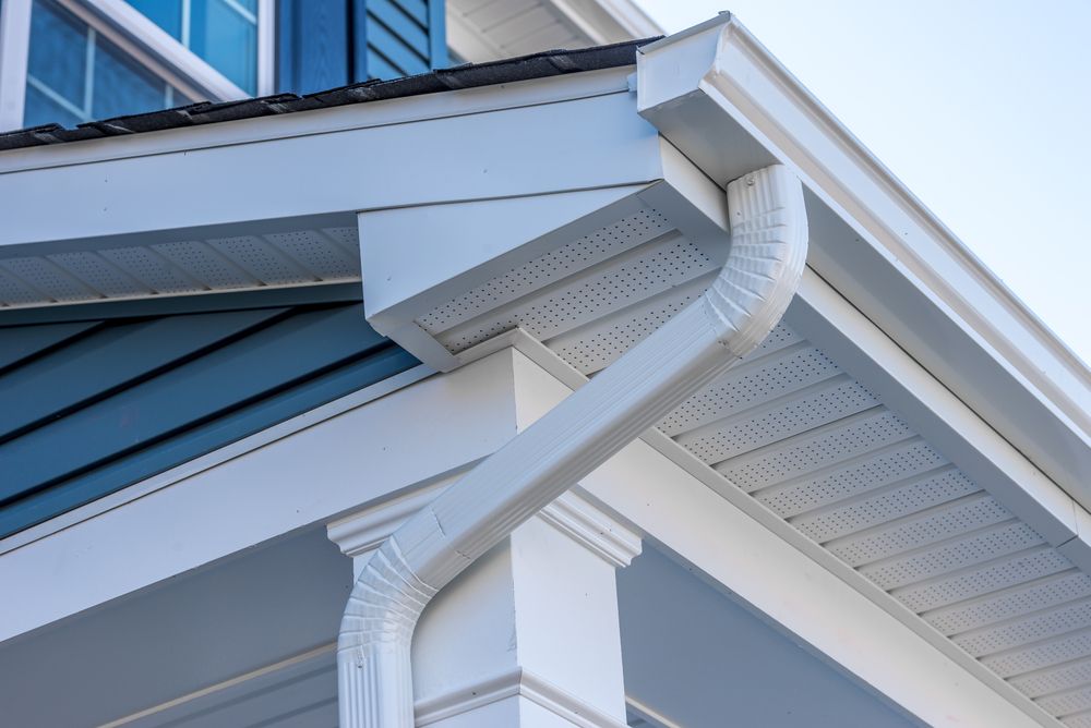 White gutter and downspout on a house roof corner with blue siding and soffit vents