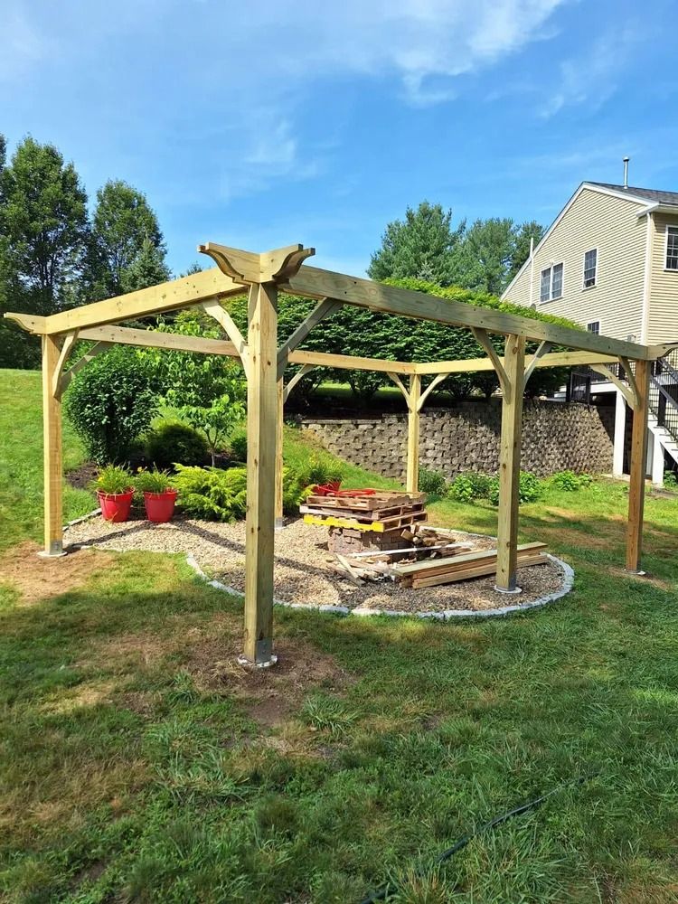 Wooden pergola shelter over a stone fire pit in a grassy backyard with potted flowers and a house nearby
