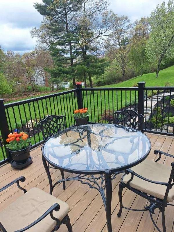 Patio table and chairs overlooking a garden and trees, with potted flowers and a black metal railing.