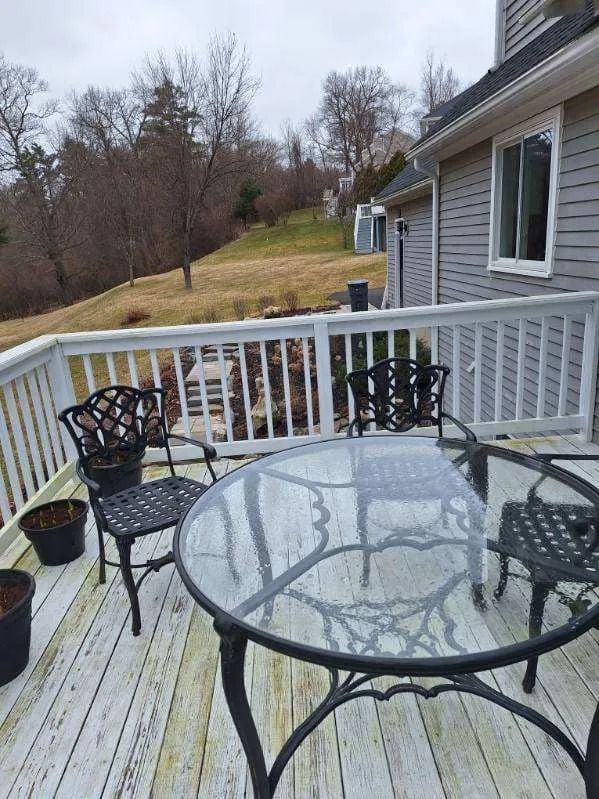 Glass patio table and black chairs on a deck beside a house, overlooking a grassy hillside.