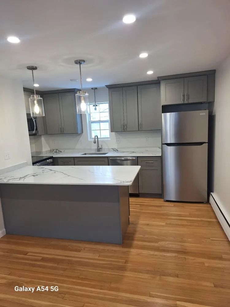 Modern kitchen with gray cabinets, white island, stainless refrigerator, and hardwood floors.