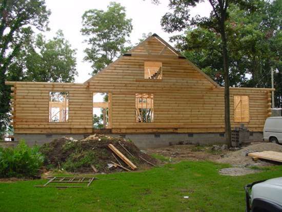 Wooden log cabin under construction, windows cut out, surrounded by grass and trees.